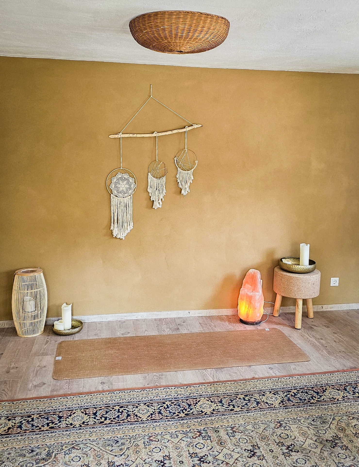 Yogaraum im LIVA Meiningen mit warmer ockerfarbener Lehmwand, handgemachten Traumfängern aus Makramee, Rattan-Deckenleuchte, Himalaya-Salzsteinlampe, Kerzen und Jute-Yogamatte auf orientalischem Teppich